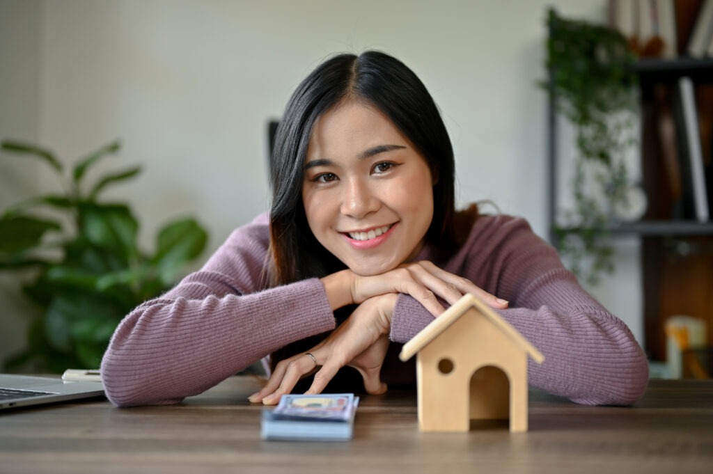 Smiling woman in a mauve sweater sits at a table with a small wooden birdhouse and playing cards in front of her indoors