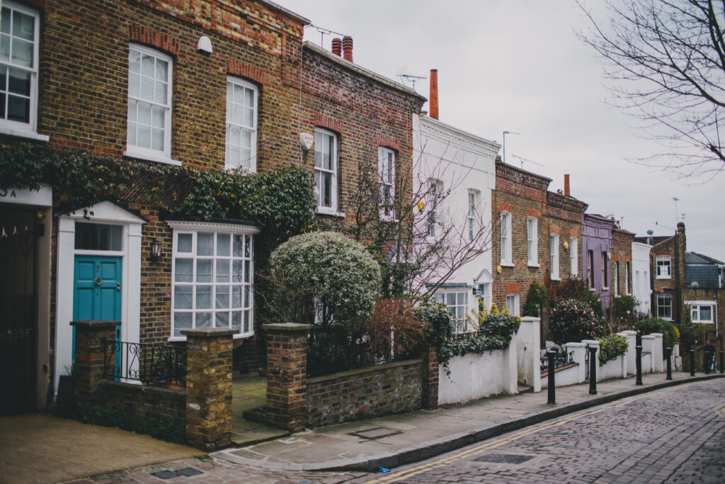 Row of brick townhouse facades with white framed windows and small front gardens on a cobblestone street under an overcast sky